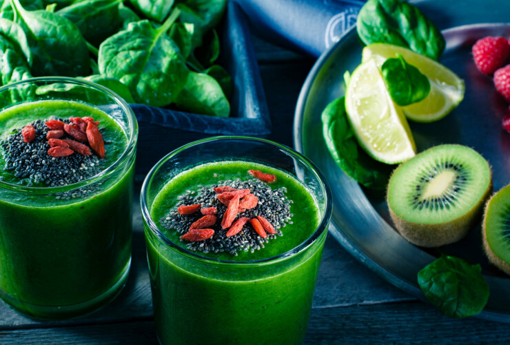 Fresh green smoothie sprinkled with chia seeds and goji berries in the background fruits and vegetables on wooden table.
